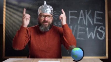 Bir adam video blogcusu arka plandaki tebeşir tahtasında Sahte Haber metniyle oturuyor. Folyo şapkalı komplo teorisyeni kameraya bakarken kendinden emin bir şekilde bir şey söylüyor..