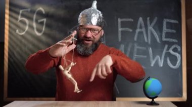 Bir adam video blogcusu arka plandaki tebeşir tahtasında Sahte Haber metniyle oturuyor. Folyo şapkalı bir komplo teorisyeni elinde tahta adam kuklası tutuyor..