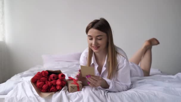 Молода красива біла жінка лежить вдома на самоті на ліжку. Букет квітів тюльпанів лежить біля коробки в обгортці для ремесел. Візьміть знак і прочитайте привітання з днем народження . — стокове відео
