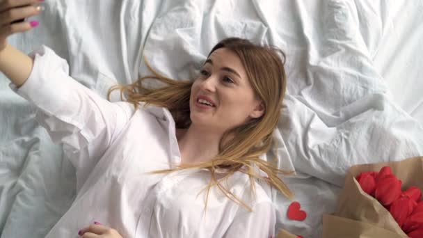 Piękna biała kobieta leży na łóżku w sypialni. Przytrzymaj smartfona, aby czat wideo online z przyjaciółmi. Kwiaty są w pobliżu. — Wideo stockowe