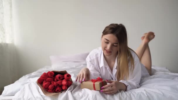Ευτυχισμένο λευκό κορίτσι ξαπλώνει στο κρεβάτι με το στομάχι της και εξετάζει το δώρο της και ένα μπουκέτο λουλούδια από κόκκινες τουλίπες από τον αγαπημένο της άντρα. Διακοπές γυναικών και μια έκπληξη για τη σύζυγο. — Αρχείο Βίντεο