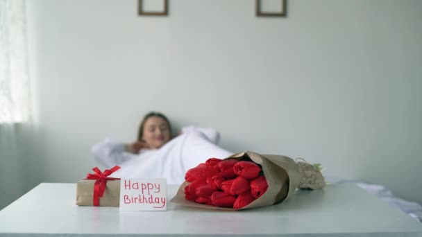 A gift and a bouquet of red tulips on a birthday near the bed of an awakened young girl on a white bed in a shirt. Happy lady in the morning in the bedroom stretches and looks at a note. — Vídeo de stock