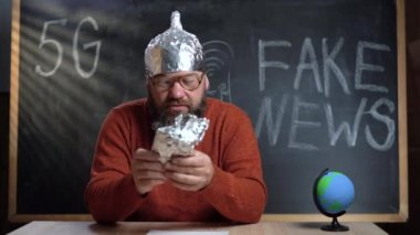 A bearded Caucasian male video blogger is broadcasting in his studio. A foil hat is worn on the head. takes out a smartphone and makes a call.