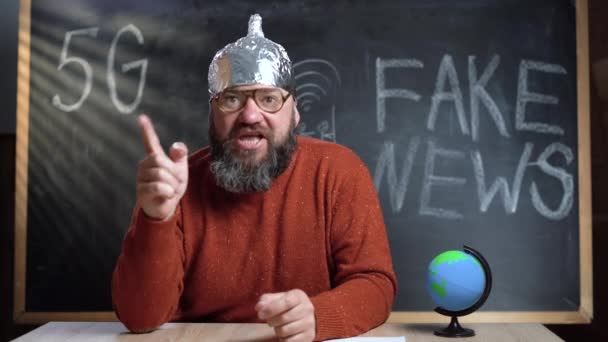 A bearded Caucasian male video blogger is broadcasting in his studio. A foil hat is worn on the head. Inadequate psycho talks about conspiracy theory. — Stock videók