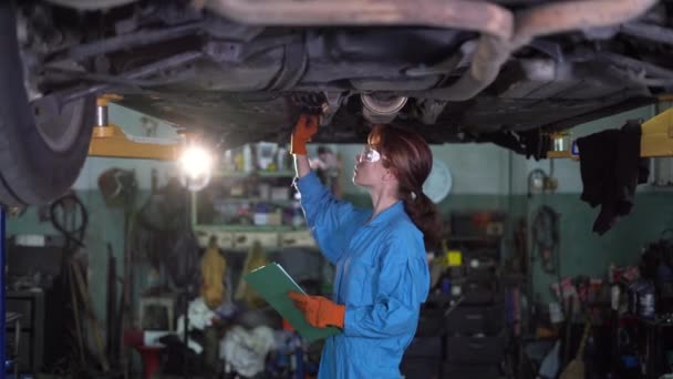 Kvinnlig bilmekaniker som jobbar under en bil i ett garage. Porträtt av en koncentrerad kvinnlig mekaniker. flickan gör en inspektion av den upphöjda bilen, skriver ner skadan på ett pappersark. — Stockvideo