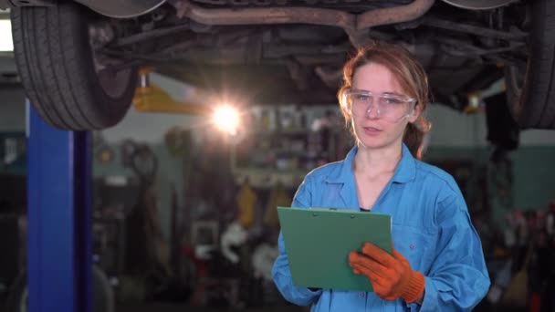 Μια γυναίκα μηχανικός στέκεται στο παρασκήνιο ενός ανυψωμένου αυτοκινήτου. κρατά το δισκίο στα χέρια του και καταγράφει τη ζημιά. Σημάδια με στυλό. Αντιγραφή χώρου. — Αρχείο Βίντεο