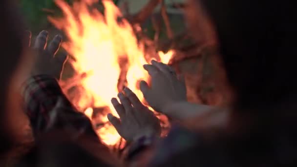 Acercamiento de dos pares de manos junto al fuego, un hombre y una mujer se sientan cerca del fuego al atardecer y se calientan. concepto de recreación al aire libre. — Vídeo de stock