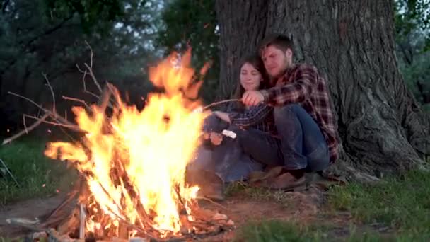 Joven pareja amorosa de turistas descansando junto al fuego en la naturaleza. Hombre guapo y mujer bonita fríen malvaviscos en una fogata cerca de la tienda en el campamento. Concéntrate en fuego. — Vídeos de Stock