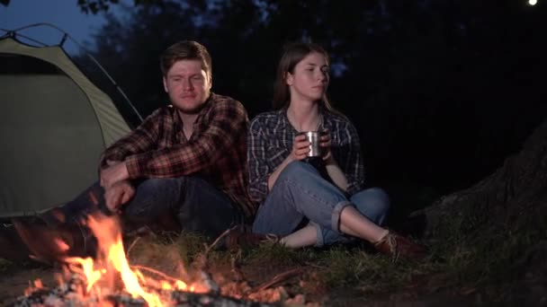 Kamperen. Een man en een vrouw zitten 's nachts bij het vuur en drinken thee of koffie uit metalen kopjes. Geliefden in de avond op de camping. — Stockvideo