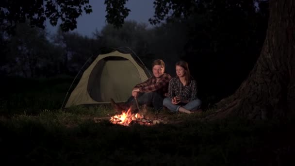 Camping. En man och en kvinna sitter vid brasan på natten och dricker te eller kaffe ur metallmuggar. Passionerad kyss i naturen. — Stockvideo