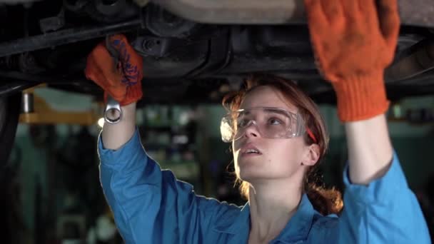 車のサービスで車の下で働く女性整備士の肖像画。手袋を着用し、車の下のキーを使用して女性に電力を供給する。本物の車の修理店. — ストック動画