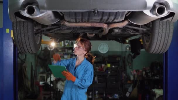 Mecânico de automóveis feminino trabalhando sob um carro em uma garagem. Capacitando a mulher usando luvas e fazendo uma inspeção de carro. Senhora trabalhando em um carro em sua oficina. — Vídeo de Stock