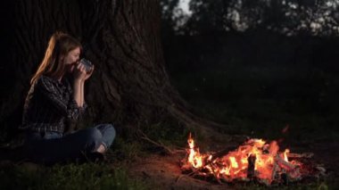 Yalnız genç bir kadın gece ormanında ateşin yanında oturmuş güneşleniyor. Yazın genç bir kız gömleğiyle doğada rahatlıyor. Metal bardaktan çay içer..