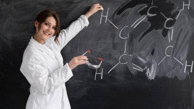 Genç bir kadın, kimya öğretmeni ya da özel öğretmen, tahtaya yakın beyaz bir önlüğün içinde duruyor ve bir formül yazıyor. Okul konseptine geri dönelim. Boşluğu kopyala.