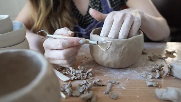 Un'artigiana siede a un tavolo in un laboratorio di ceramica e lavora su una ciotola di terra. Con l'aiuto di uno strumento speciale, raschia i pezzi in eccesso di argilla. Una tazza di argilla. Scultore. — Video Stock