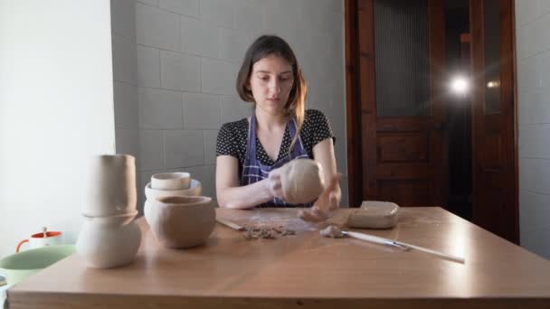 Een pottenbakker in een pottenbakkerij. Handgemaakt. Keramist. Een kopje klei. Beeldhouwer. Een jonge vrouw zit aan een tafel in haar atelier en beeldhouwt een kruik uit klei. Kopieerruimte. — Stockvideo