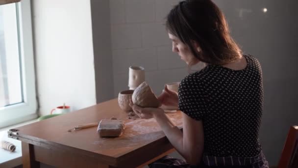 Het proces van het maken van een kan van klei. Een jonge pottenbakker zit aan tafel en maakt kleifiguurtjes. Een pottenbakker in een pottenbakkerij. Handgemaakt. Keramist. Een kopje klei. Beeldhouwer. — Stockvideo