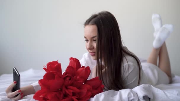 Een jong meisje ligt op het bed en neemt een selfie op haar smartphone. Een ruiker van rode tulpen ligt. — Stockvideo