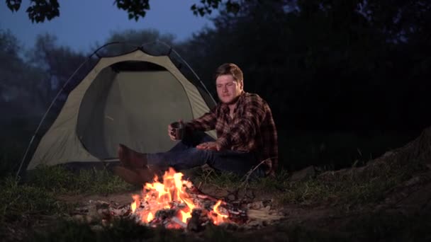 Een eenzame man zit 's nachts bij het vuur bij de tent. Drinkt thee of koffie uit een metalen mok. Het concept van outdoor recreatie weg van de drukte van de stad. — Stockvideo