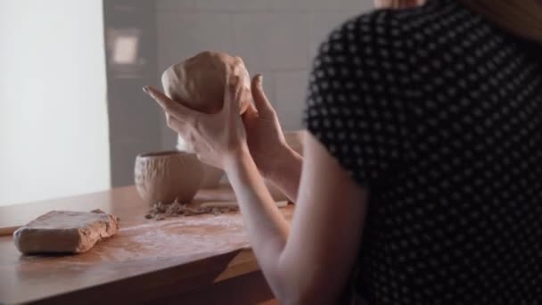 Il processo di creazione di una brocca da argilla, vista dal retro. Una giovane donna vasaio siede a tavola e fa figure di argilla. Hobby a casa nei fine settimana. scolpire una ciotola. — Video Stock