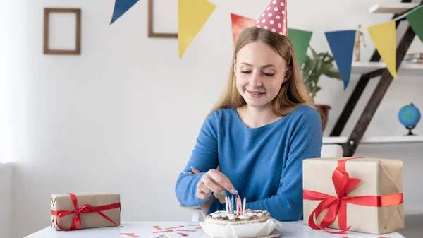 Egy üzletasszony ünnepi kalapban a születésnapján gyertyákat tesz a tortába, miközben dekorációval ül az asztalnál az irodában. Egy nő hazaérkezésének évfordulója. Boldog lány. — Stock Fotó