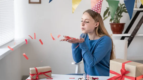 Aniversario chica sopla de confeti mientras se sienta en casa en la mesa en su cumpleaños. Fiesta en la oficina para un empleado. Un sombrero festivo en la cabeza de las chicas. Pastel y regalos. — Foto de Stock