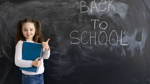 A young Caucasian girl schoolgirl stands against the background of a chalk board. The words are written back to school. holds several large notebooks in his hands and a thumb up.