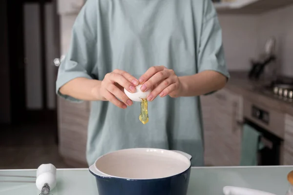 Eine Frau im T-Shirt zerbricht in der Küche ein Hühnerei und gießt das Eiweiß in einen Keramiktopf. Der Prozess der Herstellung von hausgemachten Kuchen. — Stockfoto