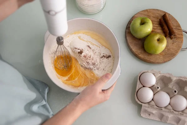 Kucharka w kuchni ugniata ciasto mikserem z trzepaczką w ceramicznym rondlu. Jabłka i mnóstwo kurzego jajka na placek. — Zdjęcie stockowe