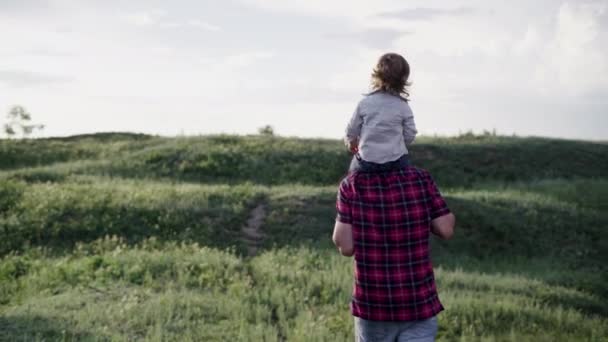 幸せな家族は、父親は牧草地を歩いて夕日を見ている彼の肩の息子を保持しています。パパと赤ちゃんが遊んでる。公園で幸せな家族が休んでいる. — ストック動画