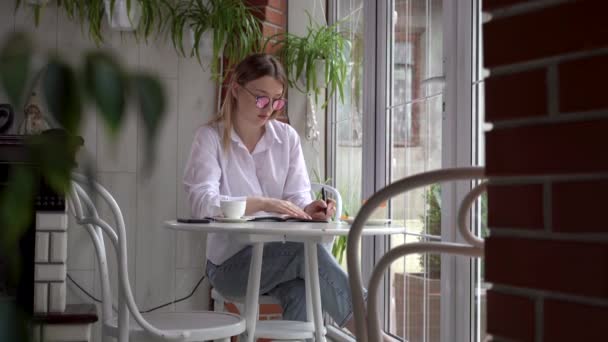 In un caffè o in un ristorante, una giovane bella signora caucasica siede a un tavolo, tiene una penna in mano e scrive qualcosa nel suo quaderno. — Video Stock