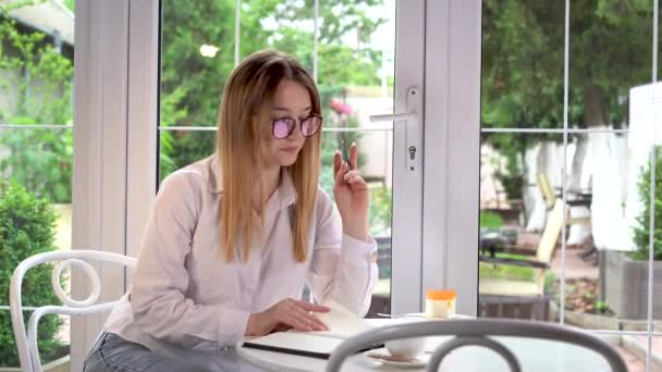 Journalistin sitzt in einem Café am Fenster und macht sich Notizen in einem Tagebuch. Junge blonde Geschäftsfrau mit Brille sitzt in einem Straßenrestaurant. Geschäftskonzept und Ideen. — Stockvideo