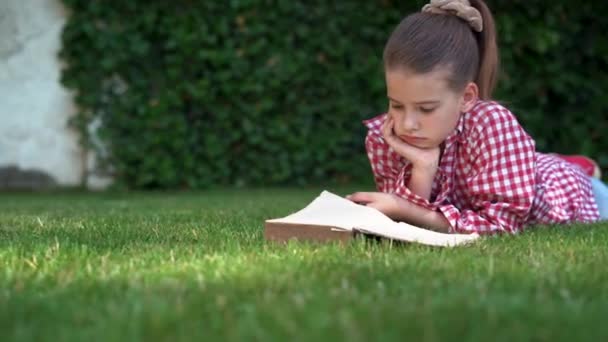 Jonge mooie blanke schoolmeisje ligt op het gazon en leest een boek. Buitenleesconcept. Huiswerk maken in het park. — Stockvideo