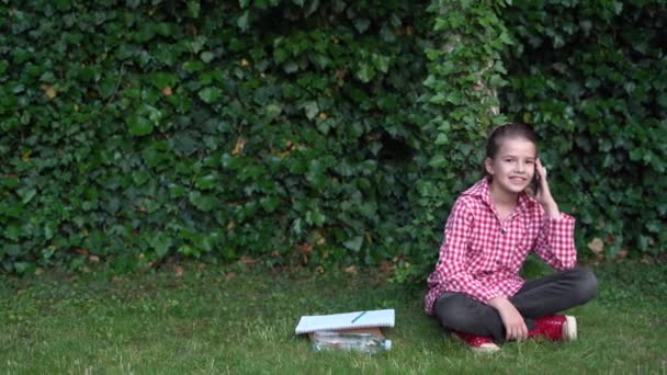 En el parque en la hierba verde una colegiala se sienta alegremente sonriendo y charlando con amigos en su teléfono inteligente. — Vídeos de Stock