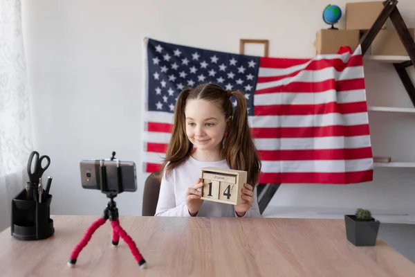 Schulmädchen sitzt zu Hause am Tisch vor dem Hintergrund der amerikanischen Flagge. hält einen hölzernen Kalender mit dem Datum des 14. Juni. Tag der amerikanischen Flagge. Videoblogger in der Nähe eines Smartphones. — Stockfoto