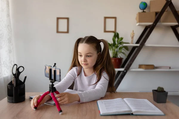 Uzaktan öğrenme çevrimiçi eğitim. Liseli kız kitap okuyor. Telefondaki video kaydı, bir tripodun üzerinde. Çevrimiçi öğrenim için bir telefon açar. — Stok fotoğraf