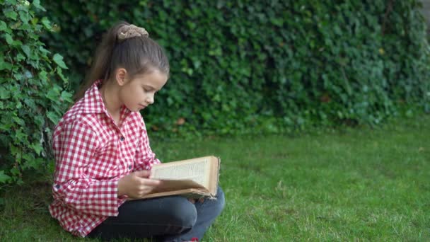 Öğrenci kız parkta çimlerin üzerinde otururken nadir bir kitap okuyor. Çocuk doğa derslerinden dinleniyor. Daha genç bir öğrenci için eğitim ve bilim. Okul çocuklarının sağlığı. — Stok video