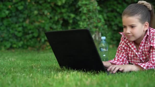 Dizüstü bilgisayarı olan genç bir kız öğrenci parkta çimlerin üzerinde yatıyor ve internette ödev yapıyor. kız öğrenci ve uzaklık öğrenimi ve açık alanda online dersler. — Stok video