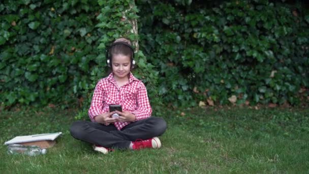 Portret van een studente die op een zonnige dag op het gazon zit met haar smartphone. Kaukasische middelbare school student meisje luisteren naar muziek met koptelefoon op de telefoon in het universiteitspark. — Stockvideo