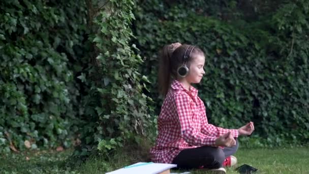 Meisje schoolmeisje in het park mediteert zitten in de lotus positie op het groene gras. het kind rust in de open lucht van studies of lessen. yoga en ontspanning concept voor schoolkinderen. — Stockvideo