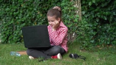 Kırmızı tişörtlü, beyaz liseli bir kız çimlerde oturur ve bir dizüstü bilgisayara bakar..