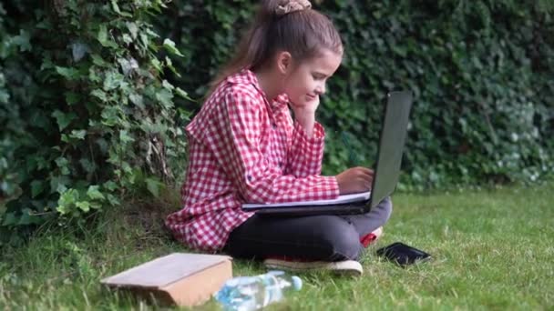 Menina branca estudante senta-se em um parque em um gramado verde com um laptop em seus pés assistindo a um vídeo e faz anotações com um lápis. conceito de treinamento ao ar livre. — Vídeo de Stock