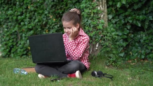 Kırmızı tişörtlü, beyaz liseli bir kız çimlerde oturur ve bir dizüstü bilgisayara bakar.. — Stok video