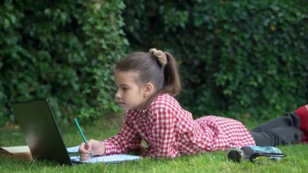 Школьница, лежащая на траве с ноутбуком, делает домашнее задание онлайн, записывая его в блокнот. Молодой студент самостоятельно учится в парке или саду. — стоковое видео