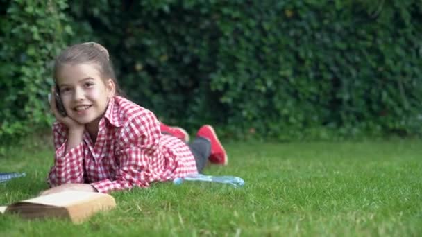 Jong lachend meisje aan de telefoon ligt in het park op het groene gras en leest een boek. Het kind rust en ontvangt audioverbinding op een smartphone. 5g — Stockvideo