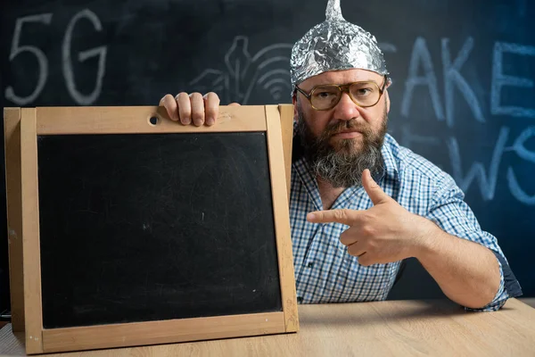 A strange bearded conspiracy theorist in a protective aluminum foil hat and glasses sits at a table and points his finger at a blank board. Fake news concept. 5g. Place for your text.