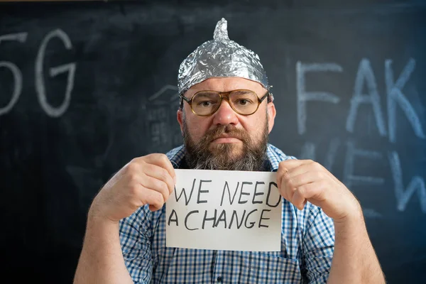 a bearded conspiracy theorist in glasses with a sign we need changes in his hands sits at the table. Fake news and conspiracy theory. Protective foil cap against magnetic radiation.