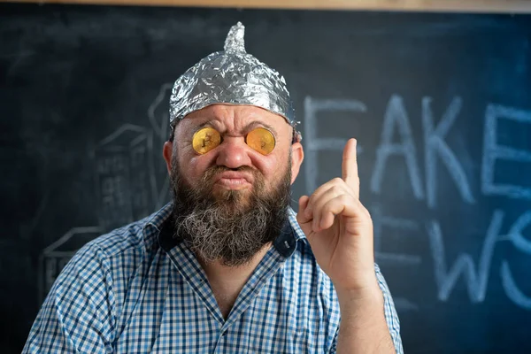 Close-up a conspiracy theorist in a protective foil hat with bitcoin coins in his eyes raised his finger up on the board. Conspiracy theory concept of fake news and alternative facts. — Stock Photo, Image