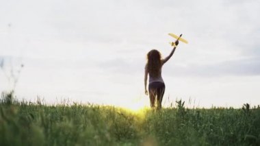 Elinde oyuncak bir uçakla sahada yürüyen güzel vücutlu genç bir kadının arka görüntüsü. Batan güneş, yumuşak odaklanma.