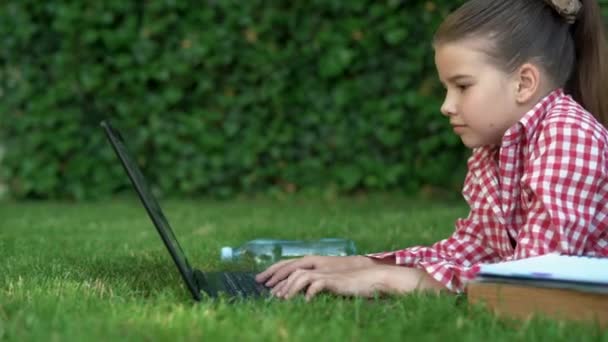 Jeune fille caucasienne se trouve sur la pelouse et travaille à l'ordinateur portable. Technologie et wi-fi, le concept de l'enseignement à distance dans la nature. — Video